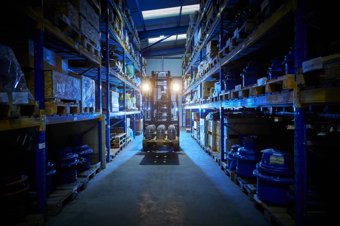 A forklift with headlights on moves down an aisle in a warehouse with shelves of industrial equipment.