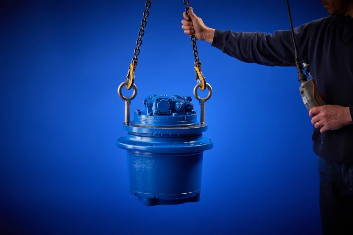 A person operates a hoist to lift a large blue industrial gear reducer against a blue background.