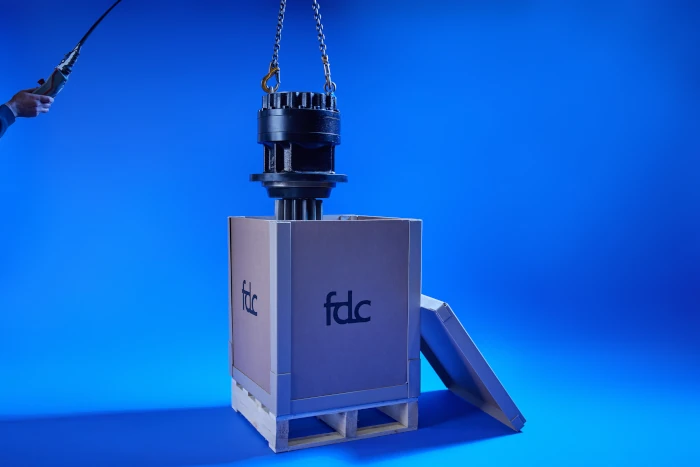 A mechanical part is being lifted by a chain above a wooden crate labeled fdc against a blue background.