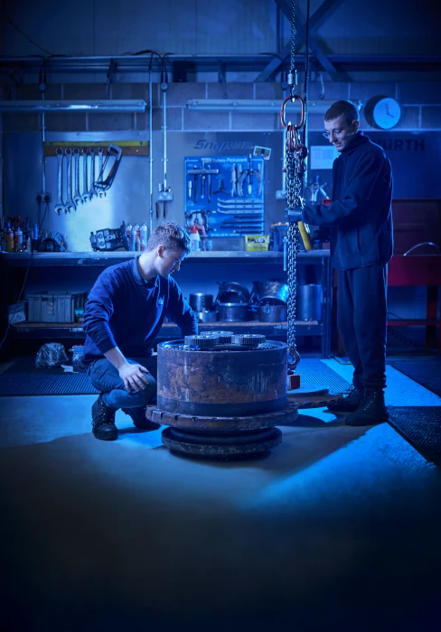 Two men work in a workshop, using a chain hoist to lift a large metal component on the floor.