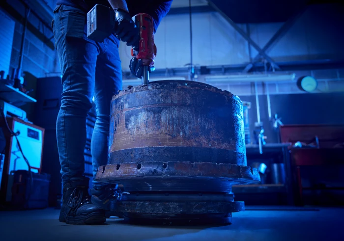 Person using a power tool on a large, rusty metal cylinder in an industrial workshop.