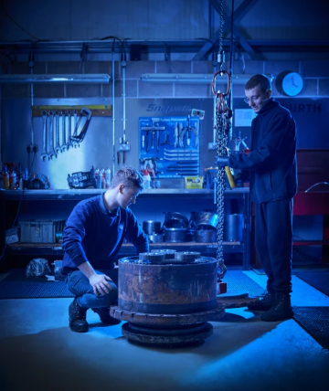 Two men work in a workshop, using a chain hoist to lift a large metal component on the floor.